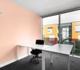 Spacious office with large windows providing a view, white desk, black office chair, peach-colored walls, and colorful exterior building visible through the windows.