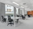 The image shows a modern, open-plan office space with large windows, white walls, and a gray carpet floor. The office is equipped with multiple workstations, each with a desk and an office chair. The desks are arranged in rows, and the space is well-lit b