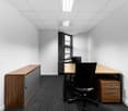 Small office space with white walls, black office chair, wooden desk, and storage cabinet. The room has a window and a suspended ceiling with lighting fixtures.