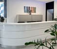 Curvaceous white reception desk, 4 chairs, gray cabinets, abstract art on wall, window, plants