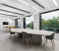 Large open office space with a long wooden conference table, gray upholstered chairs, and a wall of windows overlooking a green outdoor area. The ceiling has recessed lighting fixtures and the floor is a light-colored wood laminate. There is a TV mounted