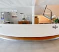 White curved reception desk, wooden desk behind with flower vase, stairs to the right, brand logo on the desk, two small staircases to the right with black rails, lamps mounted on the ceiling, white walls