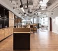 Open concept office space with wooden shelving units, black countertops, and modern lighting fixtures. The space features a kitchen area with a dishwasher and a seating area with tables and chairs.