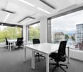 Spacious office with large windows providing a view of the city outside, white desks and black office chairs, modern lighting fixtures, and carpeted flooring.