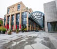 A multi-story commercial building with a glass and stone facade, featuring a large arched glass canopy entrance. The building has several balconies or terraces visible. There are red umbrellas and potted plants around the entrance area, indicating it may