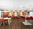 Open office space with colorful wall decor, wooden floors, round and rectangular tables, red and orange chairs, and a kitchen area with a dishwasher