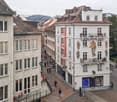 This image shows a street view of a historic city center, with a mix of old and new buildings. The buildings have a traditional European architectural style, with ornate facades and decorative elements. There are several shops and businesses visible on th