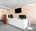 Modern office space with white reception desk, computer monitors, and a large TV mounted on the wall. The room has beige walls, a gray striped floor, and track lighting overhead.