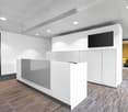 The image shows a modern and minimalist office space. The room has a reception desk with white cabinets and a gray countertop. The floor is covered with a striped carpet pattern. The walls are painted white, and the ceiling has recessed lighting. There is