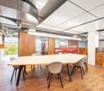 Large open office space with wooden walls, hardwood floors, and a long conference table with chairs. The ceiling has exposed ductwork and lighting fixtures. There are also built-in storage units and a window overlooking the exterior.