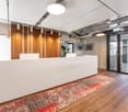 Modern office space with a large white reception desk, wooden accent walls, pendant lighting, and a patterned area rug on the hardwood floor. The space also features glass doors and windows, and appears to have a kitchen or break area with a dishwasher vi