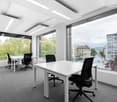 Spacious office with large windows providing a view of the city outside, white desks and black office chairs, modern lighting fixtures, and carpeted flooring.