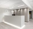 The image shows a modern and minimalist office lobby or reception area. The space features a large white reception desk with a computer monitor, pendant lighting fixtures, and a textured wall panel behind the desk. The flooring is a light-colored laminate