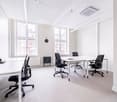 Spacious office space with large windows, white walls, and modern furniture. The room features a long table with four black office chairs, as well as individual desks with computers and other office equipment. The space appears to be well-lit and designed