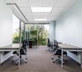 Spacious office with large windows overlooking a green outdoor area, modern furniture including desks and chairs, recessed lighting in the ceiling, and carpeted flooring.