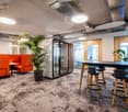 Open office space with modern furniture, including orange sofas, a large conference table with stools, and various potted plants. The space has a contemporary and relaxed feel, with exposed ceilings and ductwork, and large windows providing natural light.