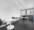 Spacious office room with white walls, large windows, black office chairs, a round white table, and a desk with a computer setup.