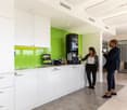Modern, open-plan kitchen with white cabinets, green backsplash, and a coffee machine. Two people are standing in the kitchen area, suggesting it is a shared office space.