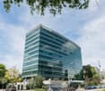A modern, multi-story office building with a glass facade and green tinted windows. The building has the Deloitte logo prominently displayed on the top. There are trees and other greenery surrounding the building, and there appears to be a parking area in