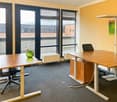 The image shows a modern office space with large windows, providing a view of the exterior. The room has a wooden desk with a computer and a green vase on it, as well as a second desk and chairs. The walls are painted in a light yellow color, and the floo