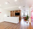 Bright and modern office space with a reception desk, wooden wall panels, and a large TV screen. The space has a clean and minimalist design with hardwood floors and potted plants. The walls have a pink gradient color scheme, adding a vibrant touch to the