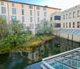 Two multi-story buildings, with windows, overlooking a canal and garden area, with trees, surrounded by greenery