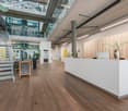 Modern and open office space with glass walls, wooden floors, a reception desk, and a welcome sign. The space features a staircase, lighting fixtures, and potted plants, creating a bright and inviting atmosphere.