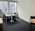 The image shows a modern office space with a desk, chairs, and a plant. The room has large windows that provide natural light and a view of the surrounding buildings. The floor is covered in dark gray carpeting, and the walls are painted a light color. Th