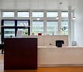 The image shows a modern office space with a reception desk, computer workstations, and large windows providing natural light. The space has a clean, minimalist design with white walls, wood floors, and black and beige furniture. The reception desk is mad