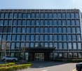 A modern multi-story office building with a glass facade, surrounded by landscaping and parking spaces in the foreground.