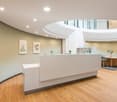 Spacious and modern office lobby with a curved white reception desk, hardwood floors, and decorative plants and artwork on the walls. The lobby has a circular skylight and a seating area with a table and chairs.