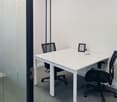 Modern office room with white desk and two black office chairs, framed picture on the desk, clear glass wall, grey carpet flooring