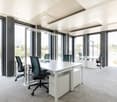 A modern office space with large windows, white desks, modern chairs, and gray carpeting.