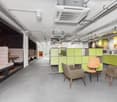 Open concept office space with modular furniture, green and gray color scheme, lounge area with chairs and tables, kitchen area with appliances including a dishwasher, exposed ceiling with HVAC system