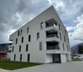 Multi-story apartment building with a modern, minimalist design. The building has a gray exterior with large windows and balconies on each floor. There is a paved walkway and landscaping in the front of the building.