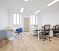 Open-space office, wooden floor, white walls, white desk, blue and beige office chairs, round table, storage cabinet, windows
