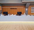 Reception desk with monitors, plants, and various items on display. Wooden wall with a large sign reading 'SPACES'. Wooden floor.
