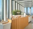 Spacious open-plan office with floor-to-ceiling windows offering a panoramic view of the city. The office features a modern, minimalist design with a wooden reception desk, potted plants, and decorative accents. The space appears to be well-lit and organi