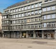 5-story building, gray exterior, multiple windows, storefronts, street sign, trees, tram tracks