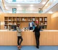 Modern office space with wooden shelves, desks, and computers. Two people are conversing at the front desk.