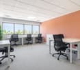 The image shows a modern office space with a bright, open layout. The room has large windows providing natural light, a suspended ceiling with recessed lighting, and gray carpeted flooring. There are several desks with black office chairs arranged in the