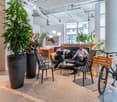 Open office space with large windows, exposed ceiling, potted plants, lounge area with couches and chairs, bicycle, and two people sitting and using a laptop