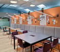 The image shows a modern and open coworking space with a mix of seating options, including tables, chairs, and couches. The walls are painted in a vibrant orange and blue color scheme, and there are several bulletin boards and whiteboards for posting info