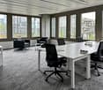 Modern office room with large windows, grey carpet, white desks, black chairs, table with various office items