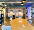 Open office space with wooden desks, blue chairs, wooden floor, wooden booths, bookshelves, flower vase, kitchen area with appliances