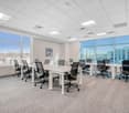 Empty office room with long white desks, grey chairs, large windows, carpet floor, and a potted plant.