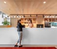 large reception desk, two women standing, modern interior, shelves with items, large glass windows, red carpet