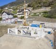 The image shows an active construction site in a mountainous, rural area. There is a large construction crane on the site, and several buildings and structures under construction. The surrounding landscape includes hills, trees, and other buildings in the