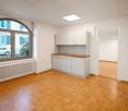 An empty room with a wooden floor, white walls, large window, kitchen cabinets, a radiator, and a wooden counter with cabinets below