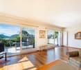 Spacious living room with large windows and sliding glass doors leading to a balcony or terrace, offering a scenic mountain and lake view. The room features hardwood floors, a sofa, and a TV stand. There is also a framed artwork on the wall.